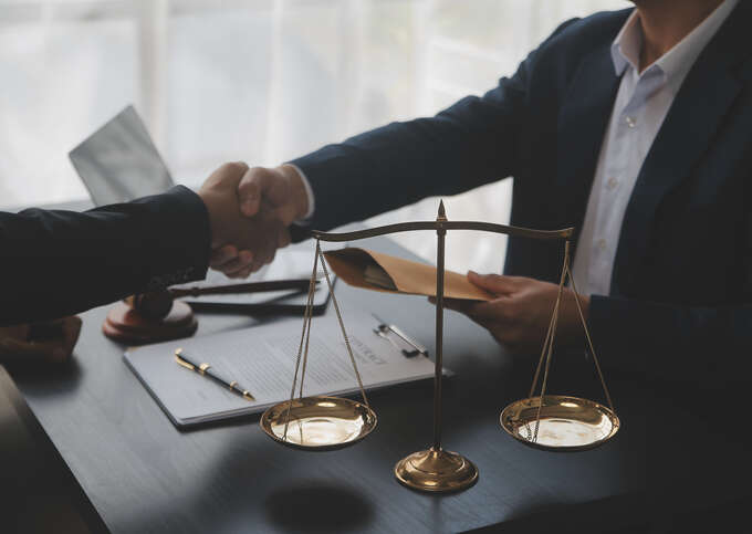 Two people dressed in suit jackets shaking hands over table with clipboard and scale