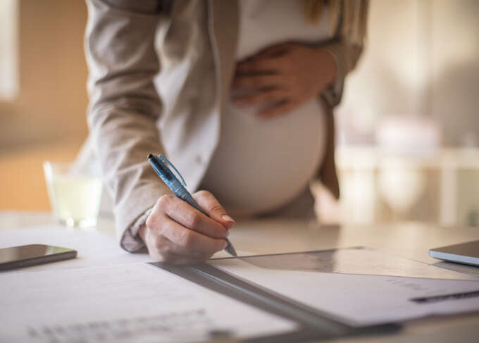 Pregnant female worker with one hand above belly and another hand writing on a document